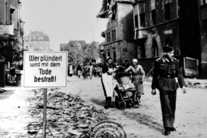 Zu sehen ist ein zerstörter Straßenzug, ein Soldat läuft vor zwei rauen, die einen Kinderwagen schieben